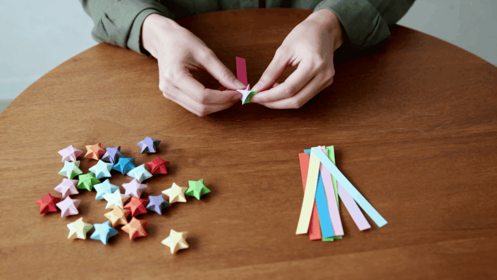 Person folding colorful paper strip into a lucky star on wooden table, with finished stars and extra paper strips neatly arranged nearby