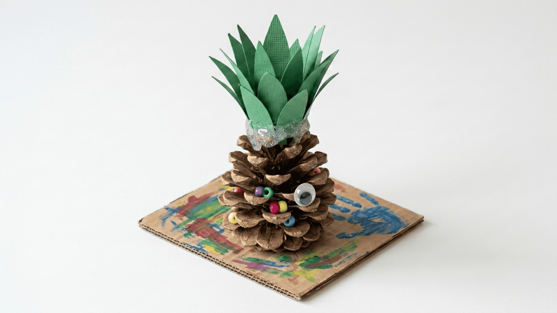 pinecone pineapple craft showing a yellow painted pinecone with green paper leaf spikes on top placed on a small paper base