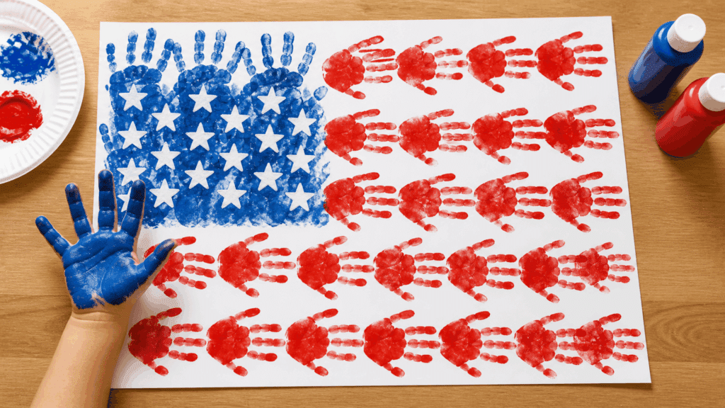 Child creating USA flag art with painted handprints in red and blue on paper, surrounded by paint bottles and craft supplies on table