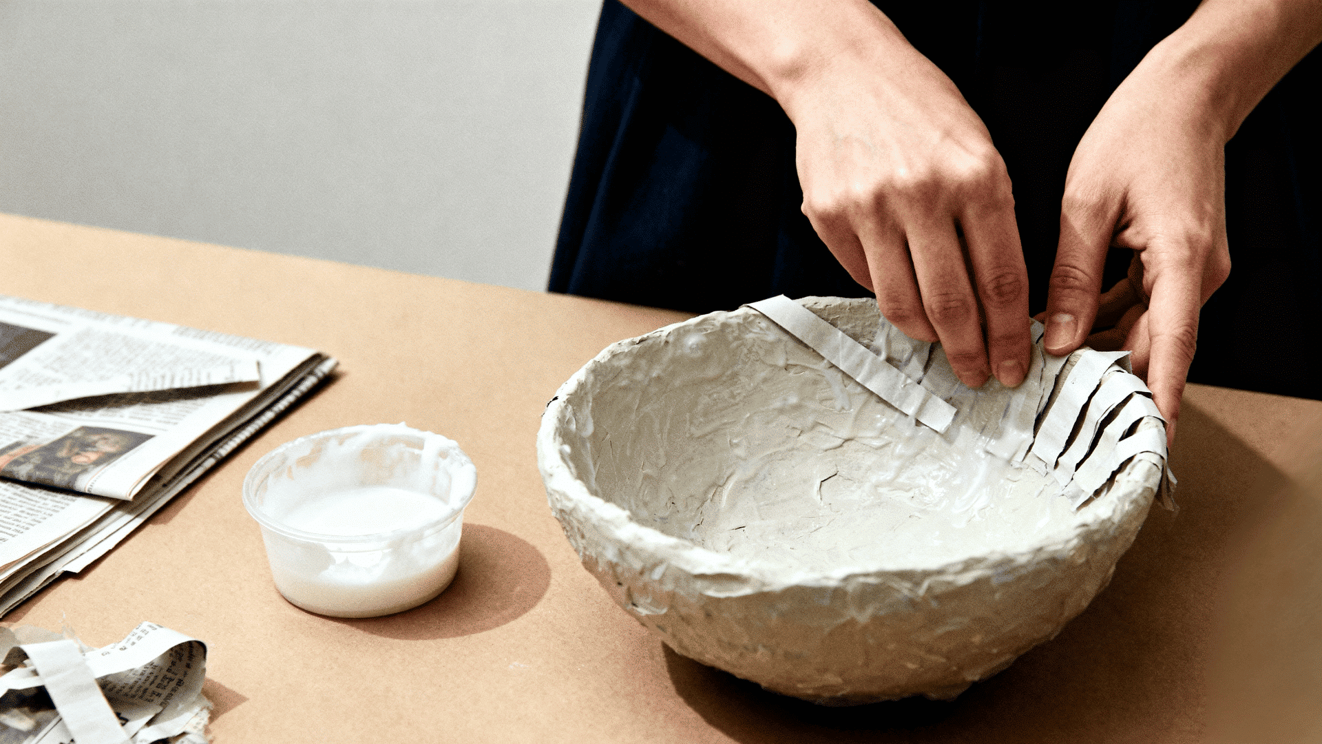 Hands crafting a paper mache bowl, applying newspaper strips with glue on a table, with tools and materials spread nearby.