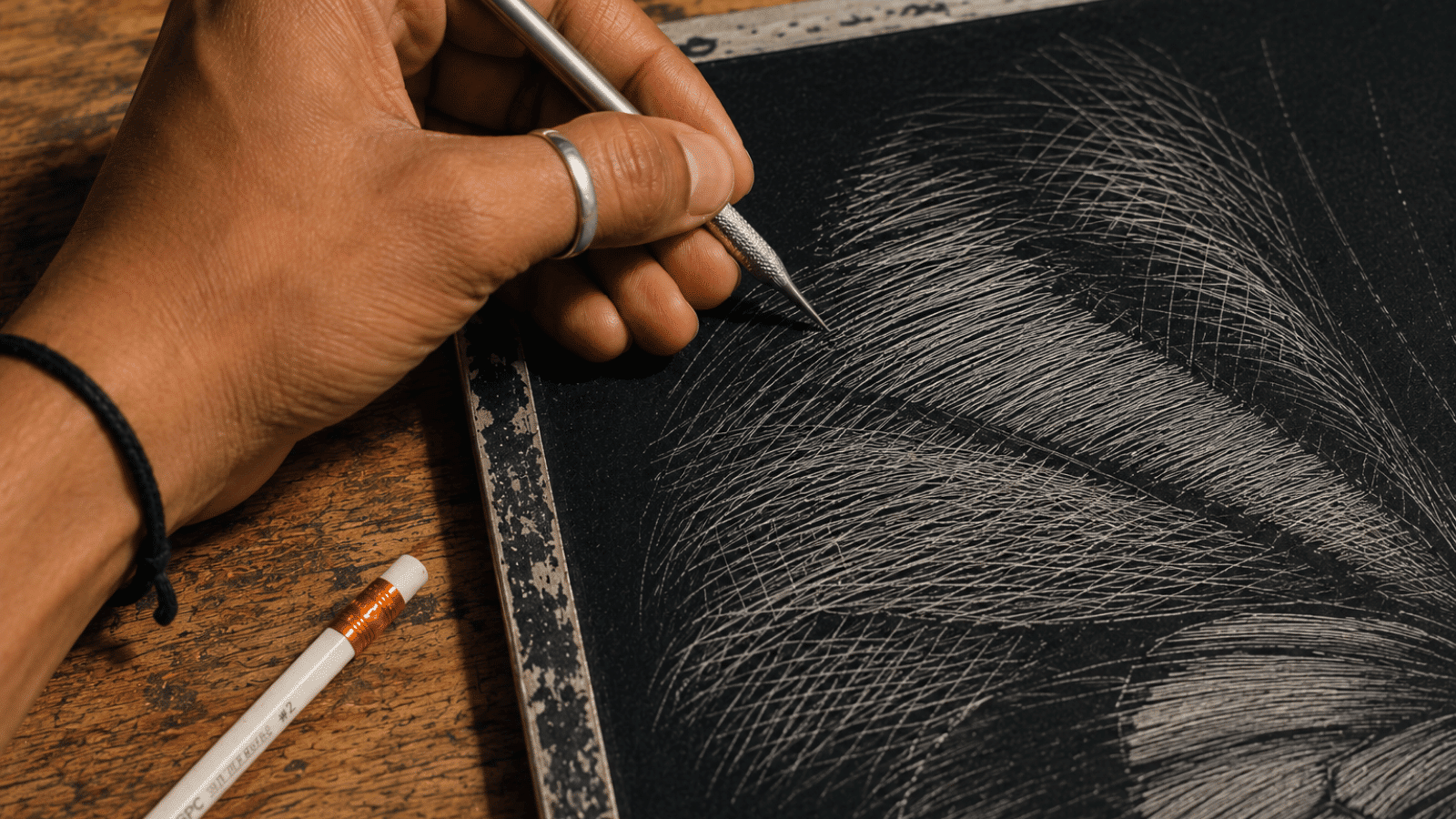 _hands scratching crossed white lines on a black scratchboard to create depth and shadow effects.