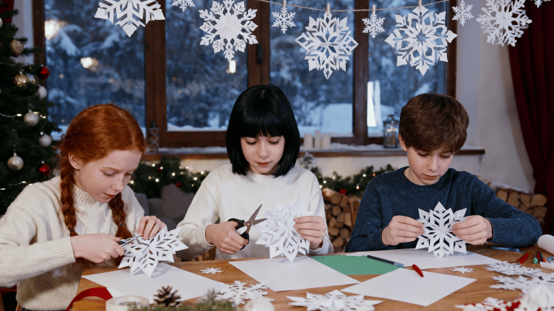 Three children cut paper snowflakes at a festive table with winter decorations and hanging snowflakes indoors tonight.