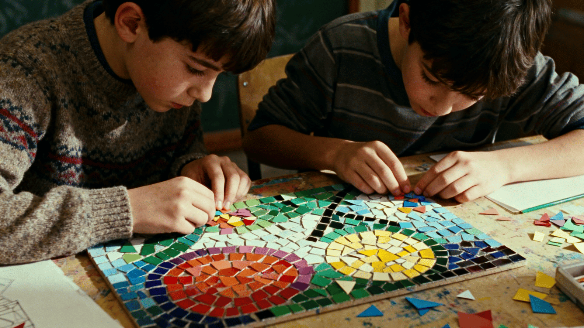 Two children carefully arrange colorful mosaic tiles on a patterned art board during a focused classroom craft activity.