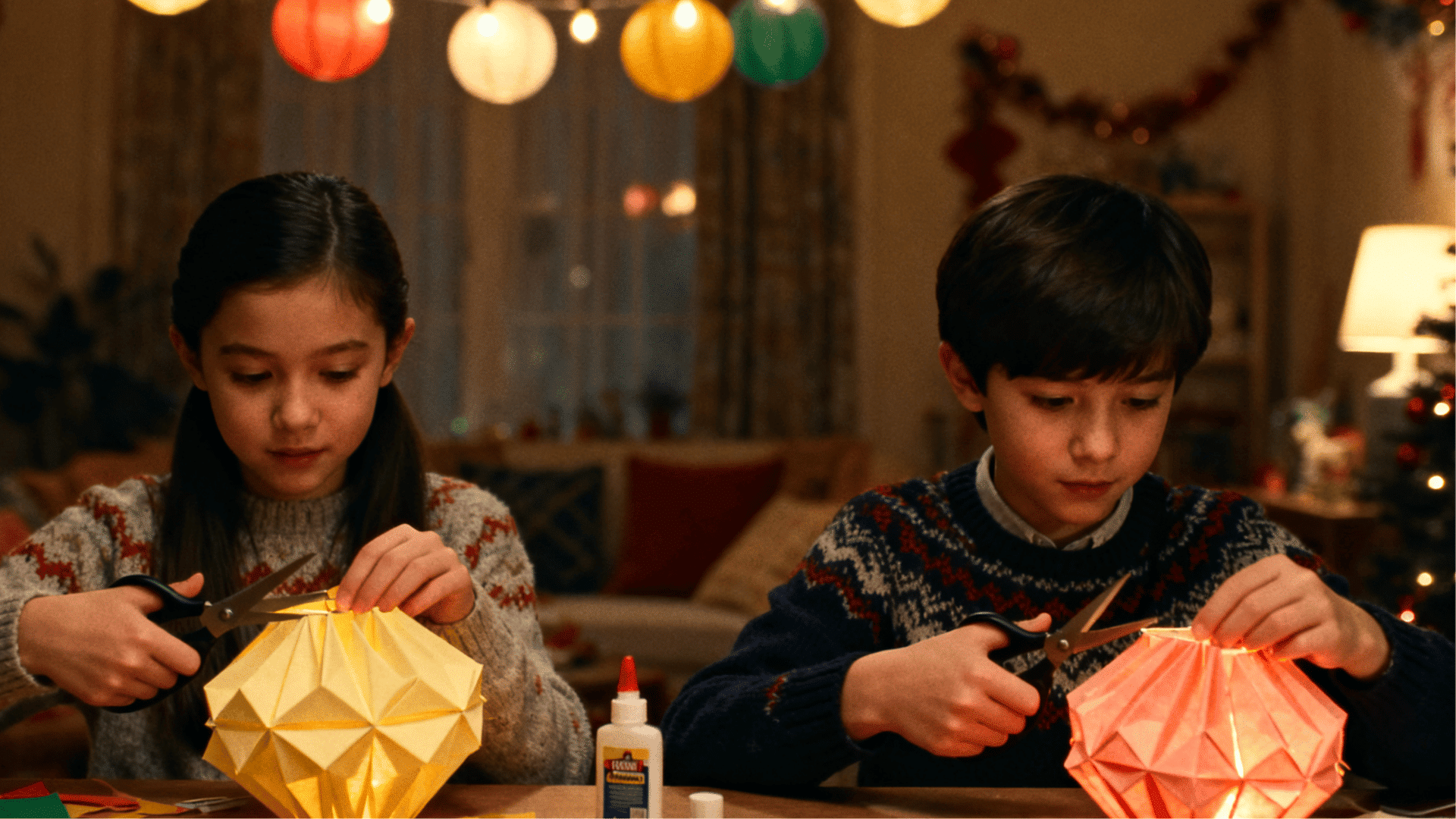 Two children create glowing paper lanterns with scissors and colorful paper during a cozy evening craft activity indoors.