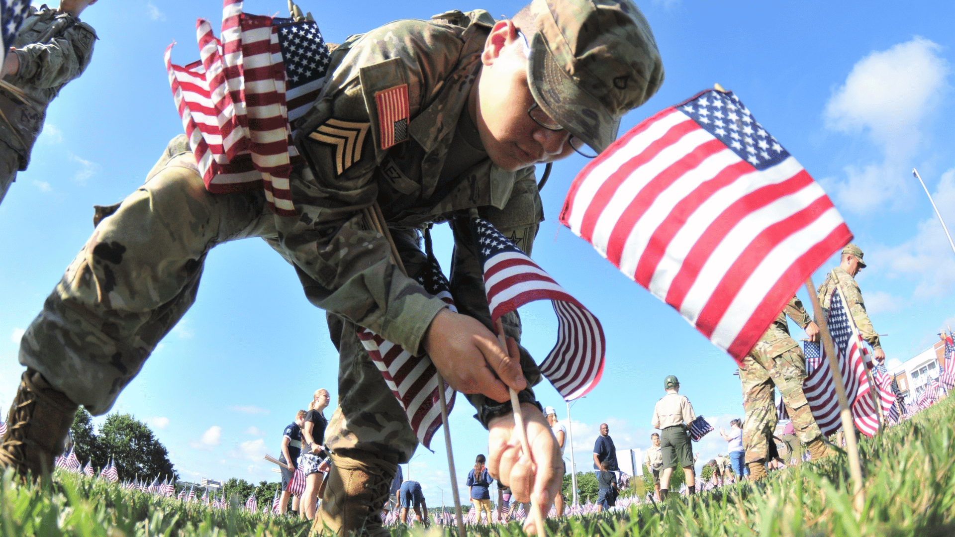 _u.s soldier plants American flags across a grassy field during a patriotic Memorial Day tribute under a bright blue sky.