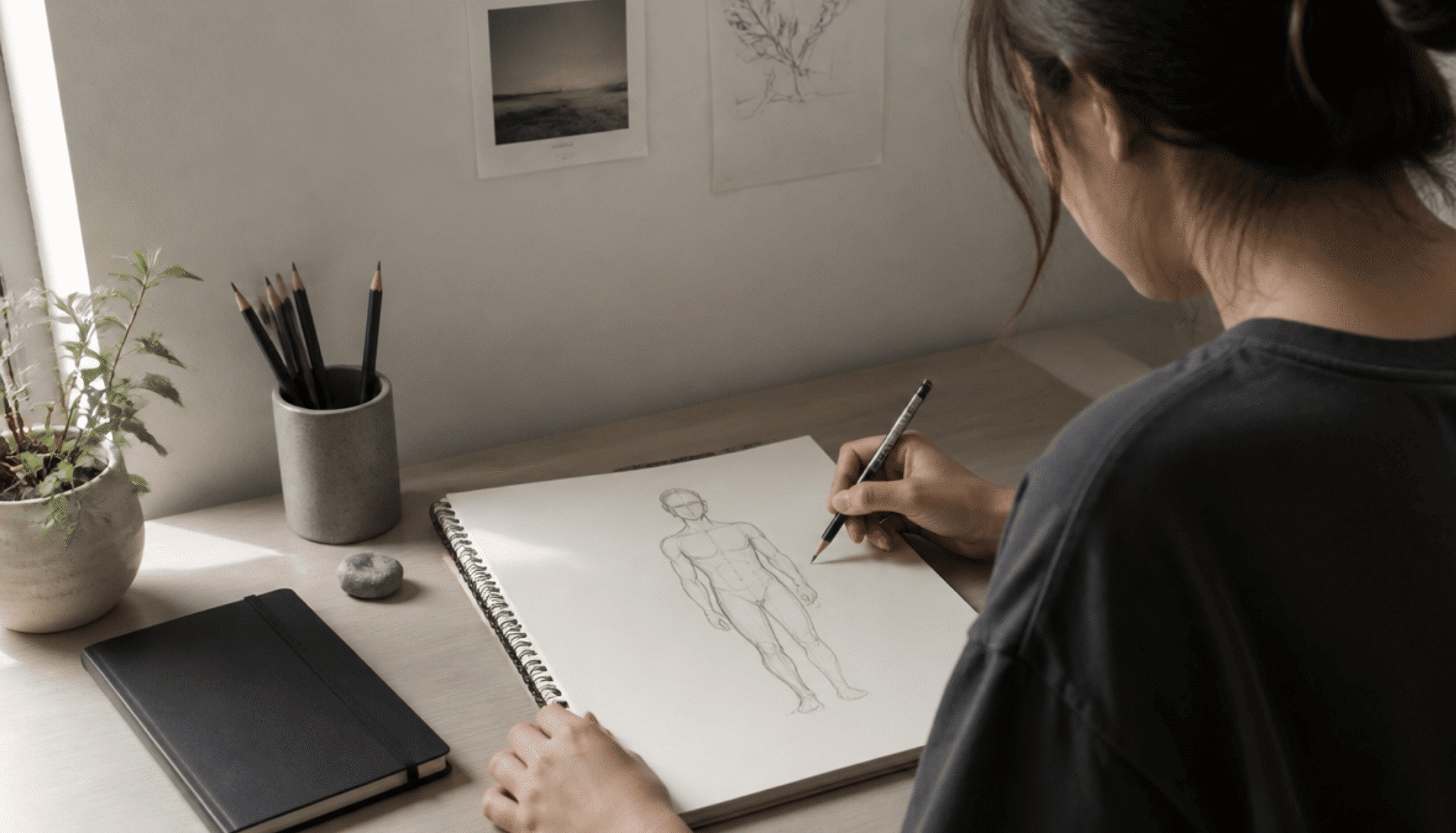 A beginner artist sketching a full human figure step by step on a sketchbook with a pencil on a clean desk