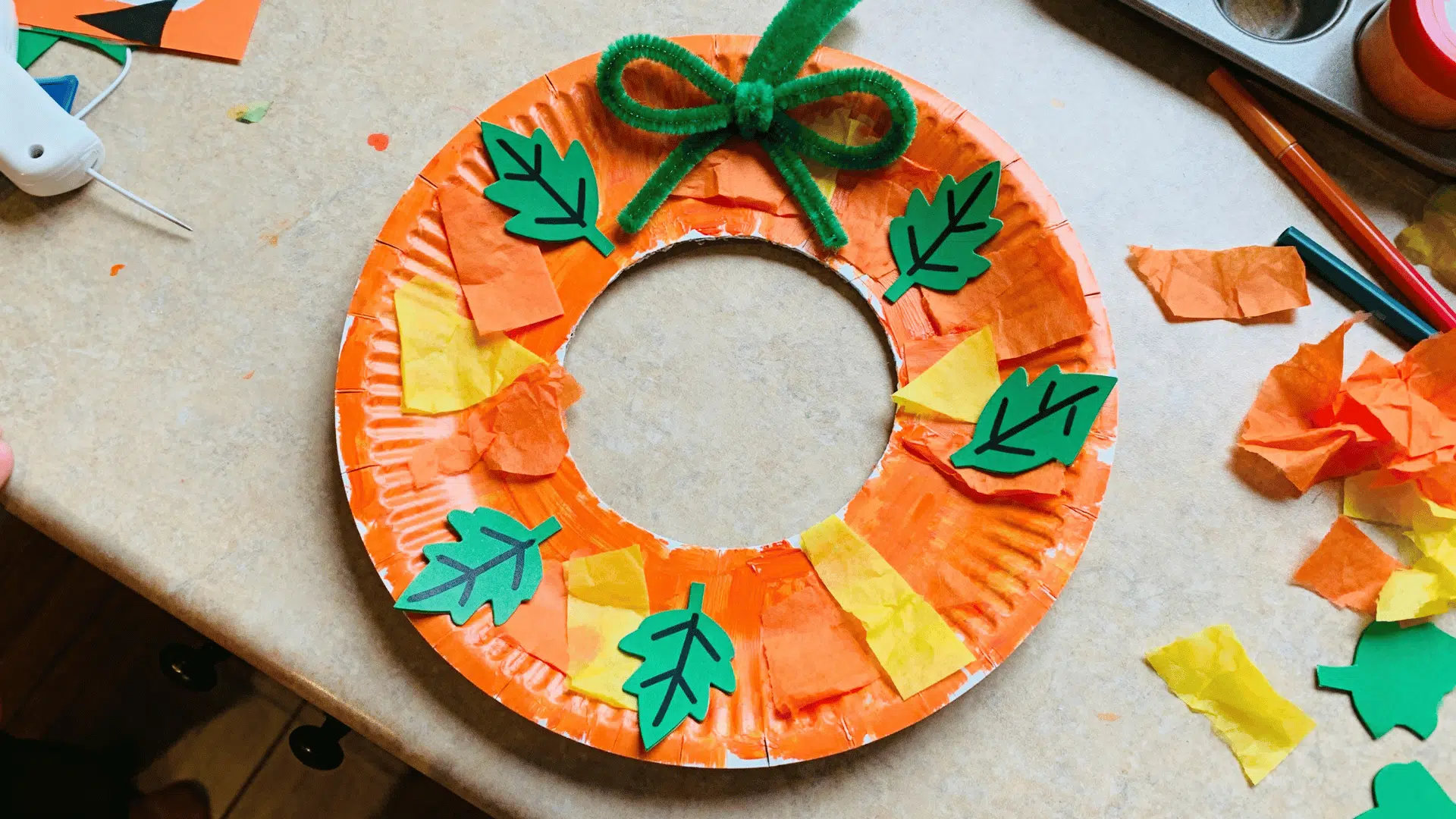 paper plate pumpkin wreath decorated with tissue paper and leaf stickers by a toddler