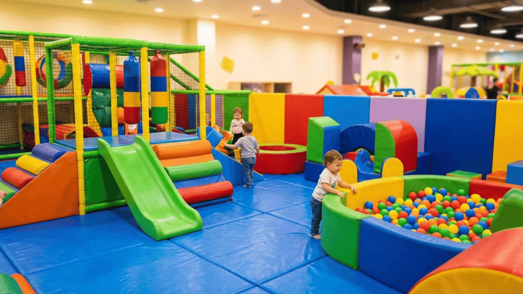 young children play among colorful soft structures, slides, and ball pits in an indoor center.