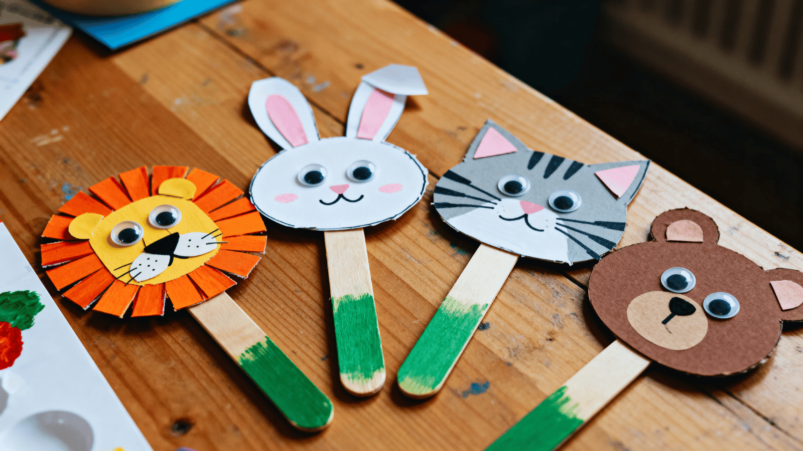 handmade popsicle stick animal faces with googly eyes and paper ears arranged on a craft table.