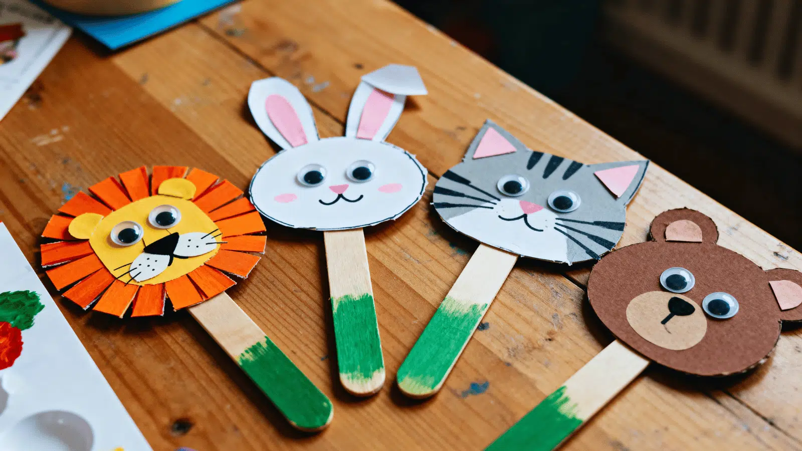 handmade popsicle stick animal faces with googly eyes and paper ears arranged on a craft table.