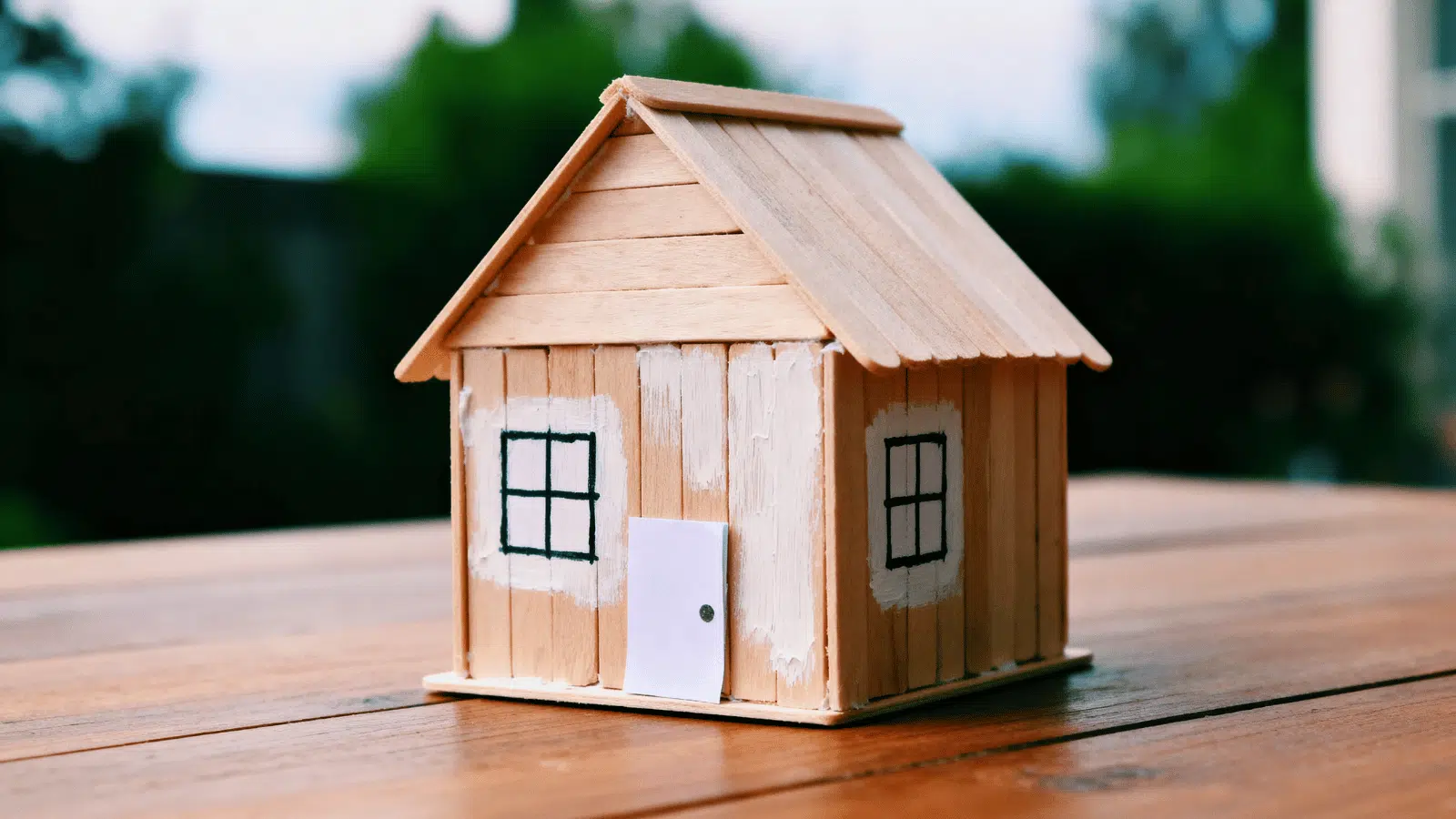 small handmade popsicle stick house with painted roof, windows, and a paper door on a tabletop.