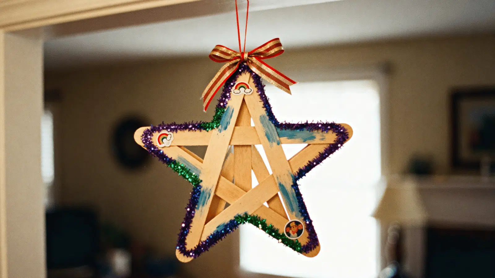 handmade popsicle stick star ornament with ribbon and glitter edges hanging indoors.