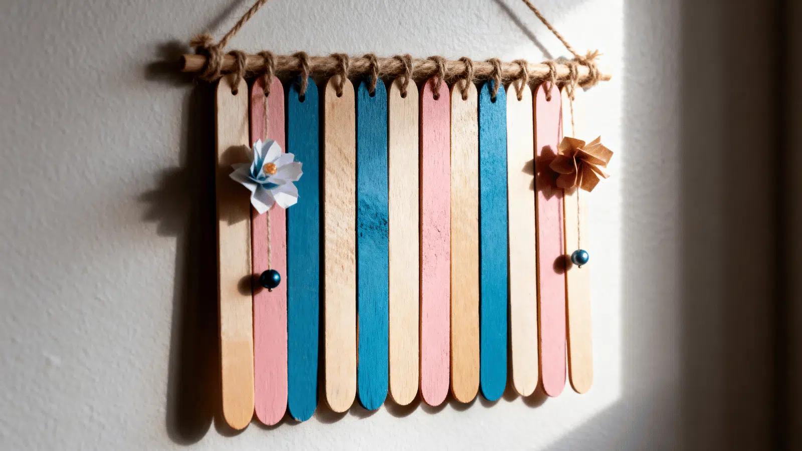 handmade popsicle stick wall hanging with beads, thread, and simple decorations on a light wall.