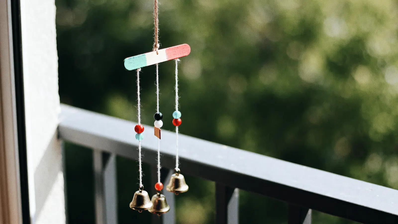 handmade popsicle stick wind chime with beads and bells hanging near a window in daylight.