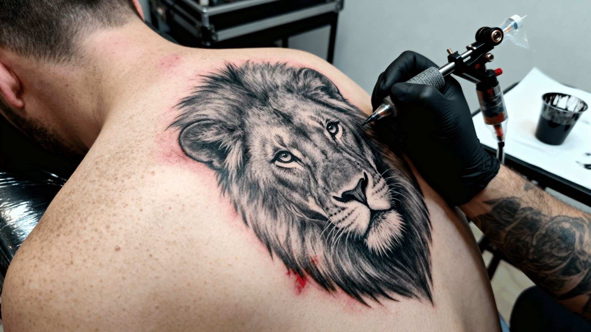 tattoo artist working on a realistic lion portrait tattoo on a person's shoulder, showing detailed shading and lifelike features.