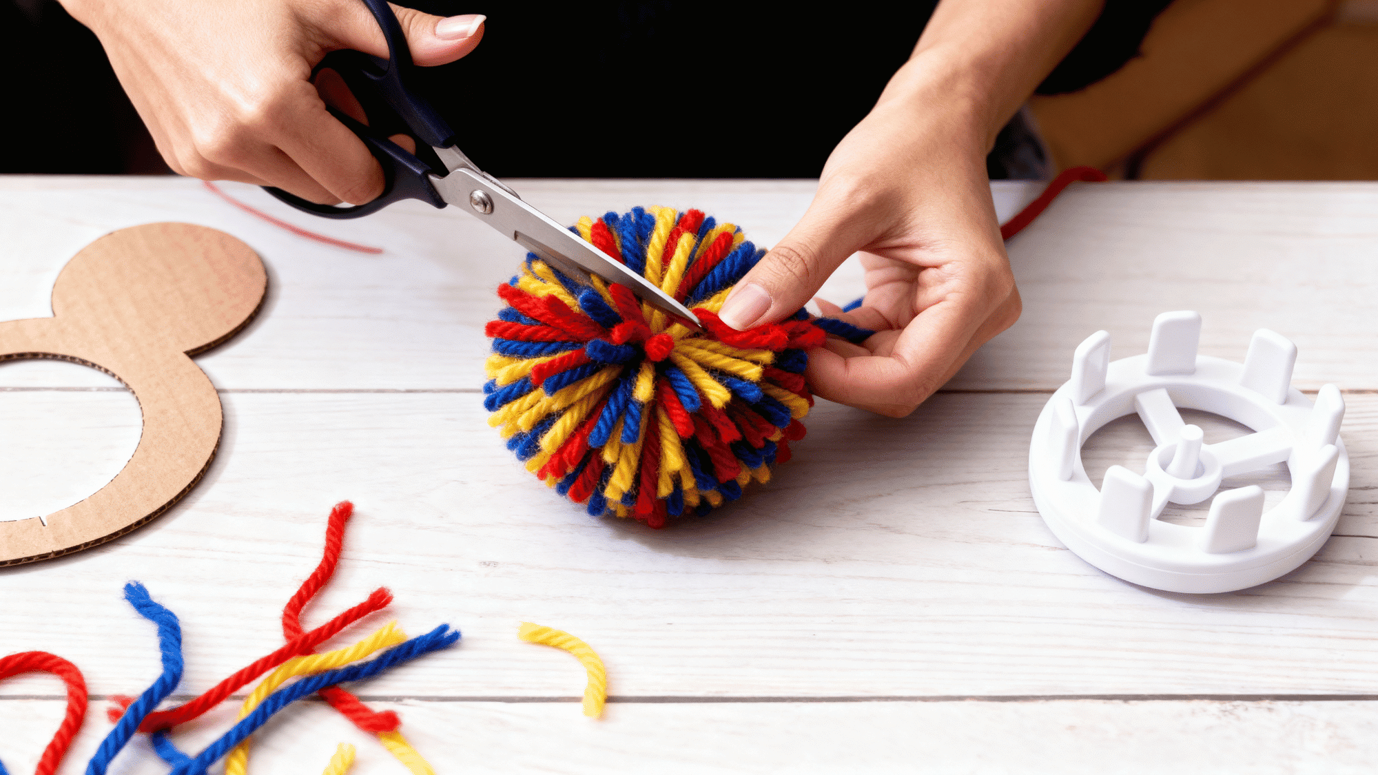 Hands trimming a yarn pom pom with scissors to create a round shape.