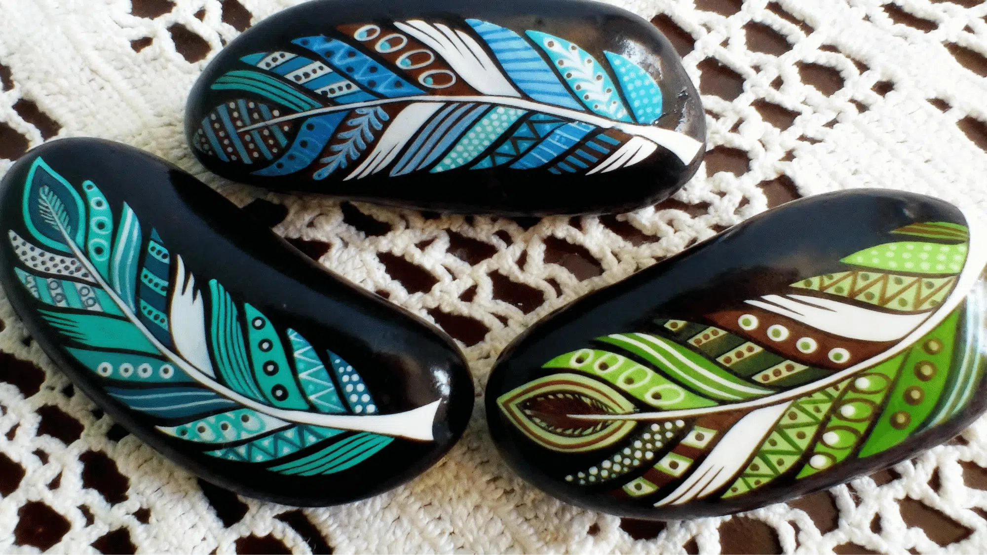 Smooth rocks painted with colorful patterned feathers in shades of blue, green, and brown, placed on a crocheted surfac