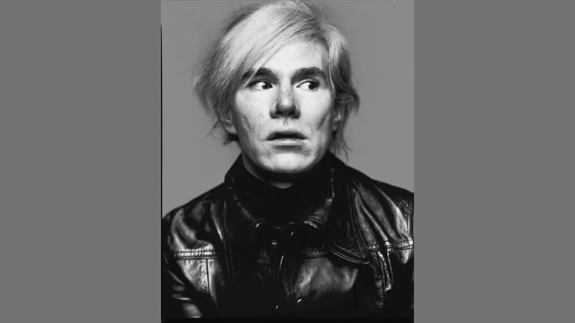 black and white portrait of artist Andy Warhol wearing a leather jacket and looking off to the side