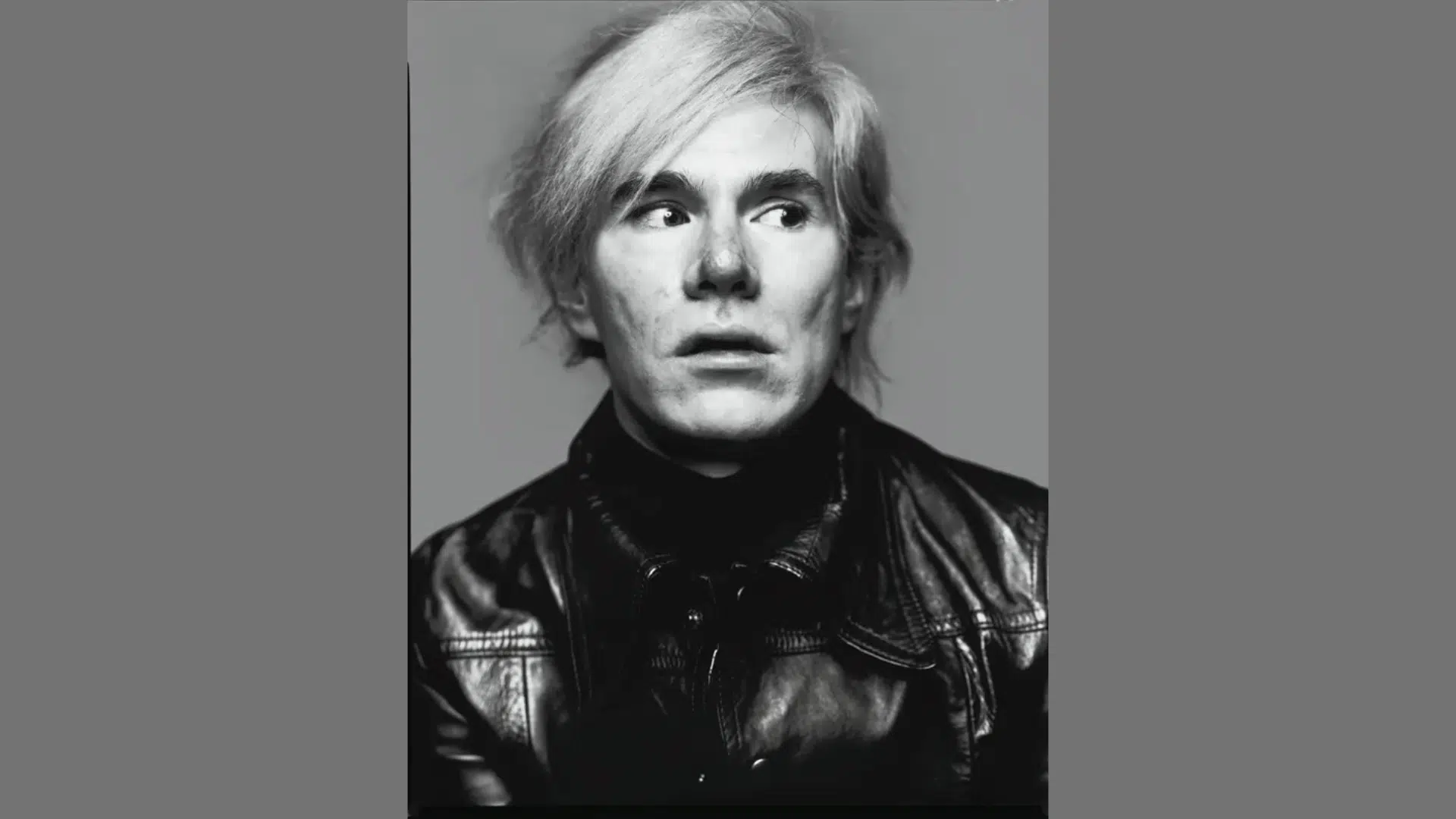 black and white portrait of artist Andy Warhol wearing a leather jacket and looking off to the side