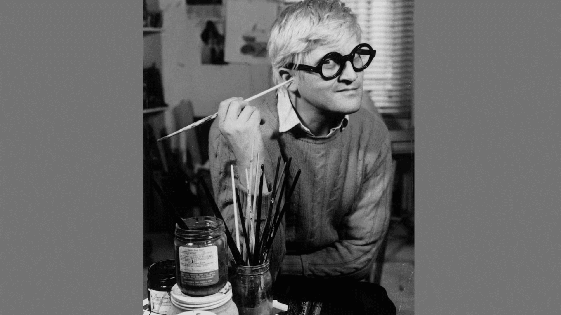 black and white portrait of artist David Hockney holding a paintbrush to his ear with jars of brushes in front