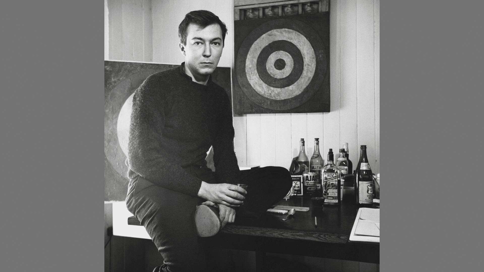 black and white portrait of artist Jasper Johns sitting on a table next to his target artwork