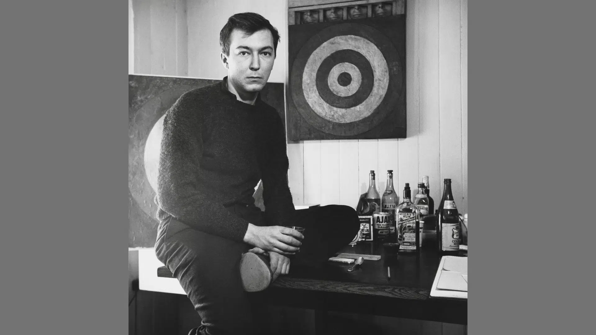 black and white portrait of artist Jasper Johns sitting on a table next to his target artwork