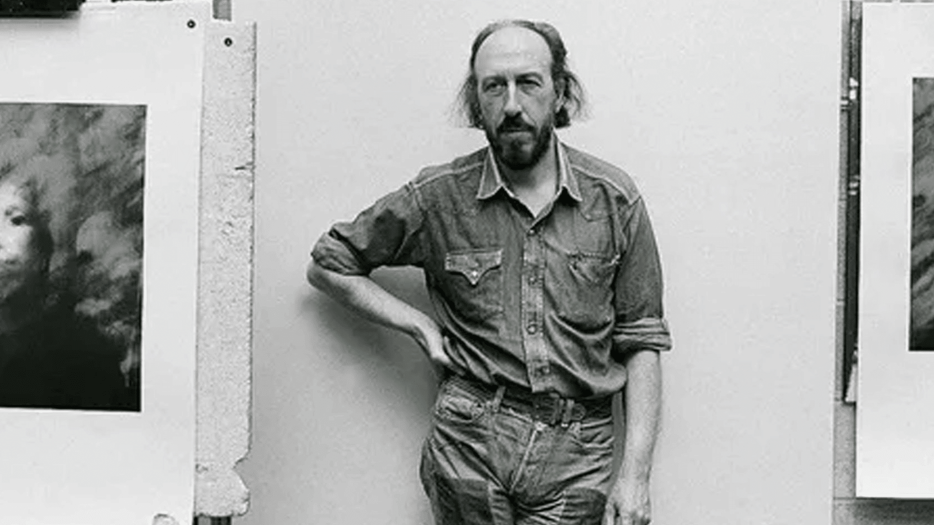 black and white portrait of artist Richard Hamilton wearing a denim shirt standing between two of his artworks