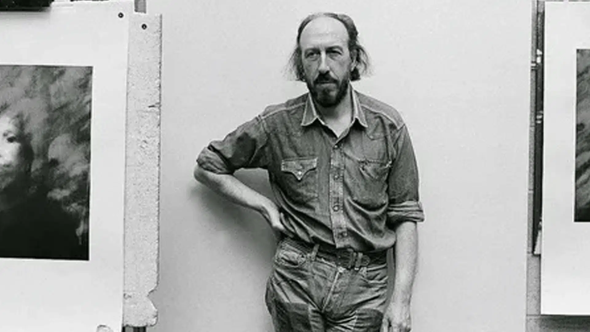 black and white portrait of artist Richard Hamilton wearing a denim shirt standing between two of his artworks