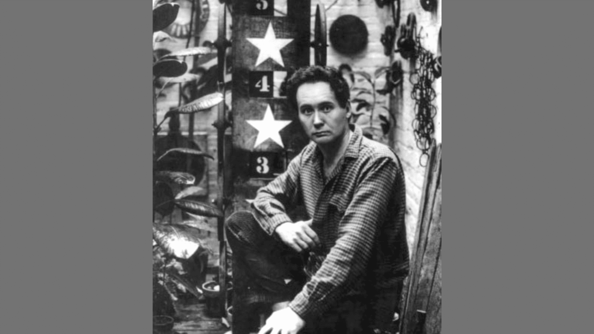black and white portrait of artist Robert Indiana sitting next to a sculpture with numbers and stars