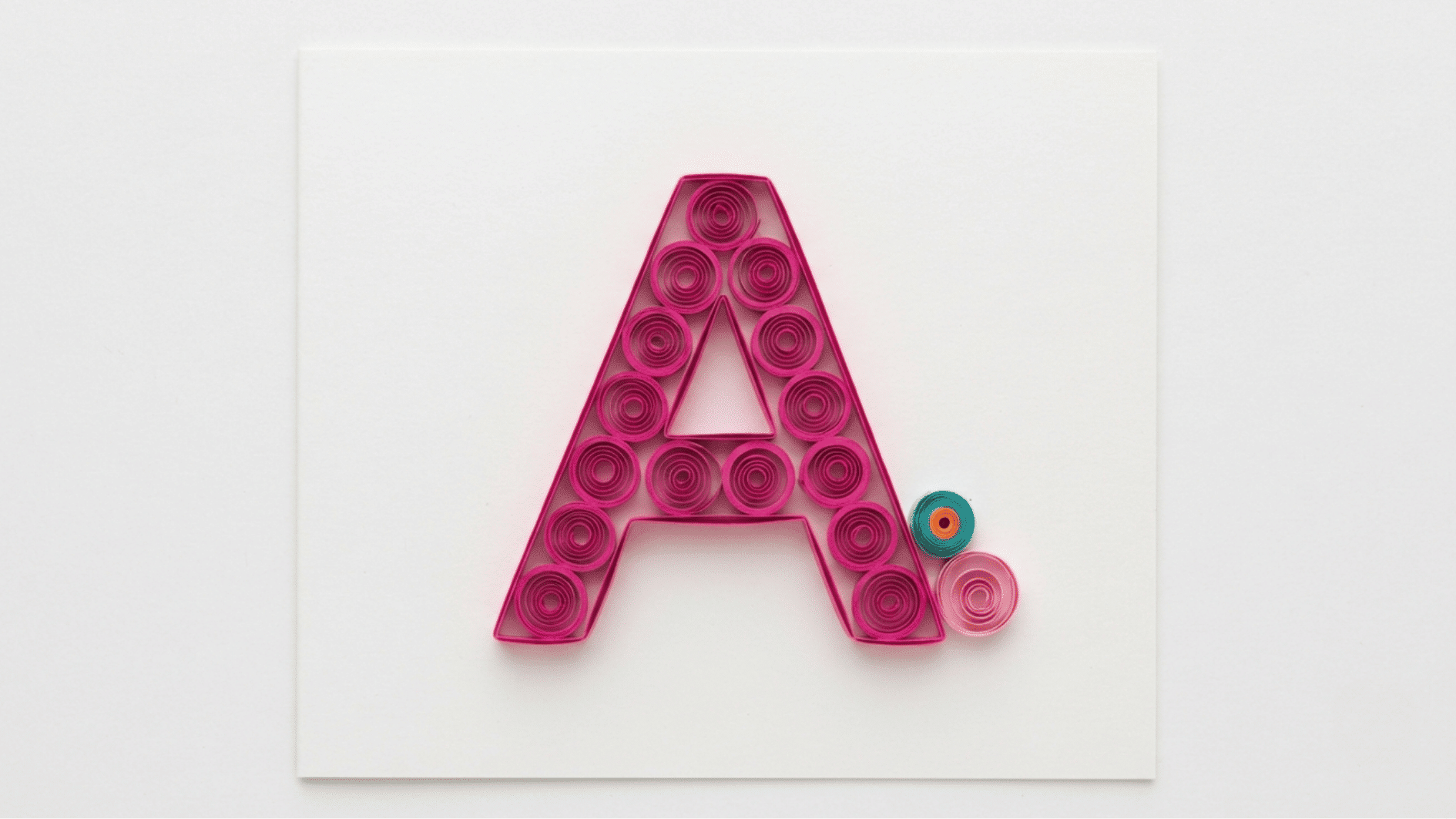 quilled paper letter a in pink coils with small teal and pink accent circles on white background