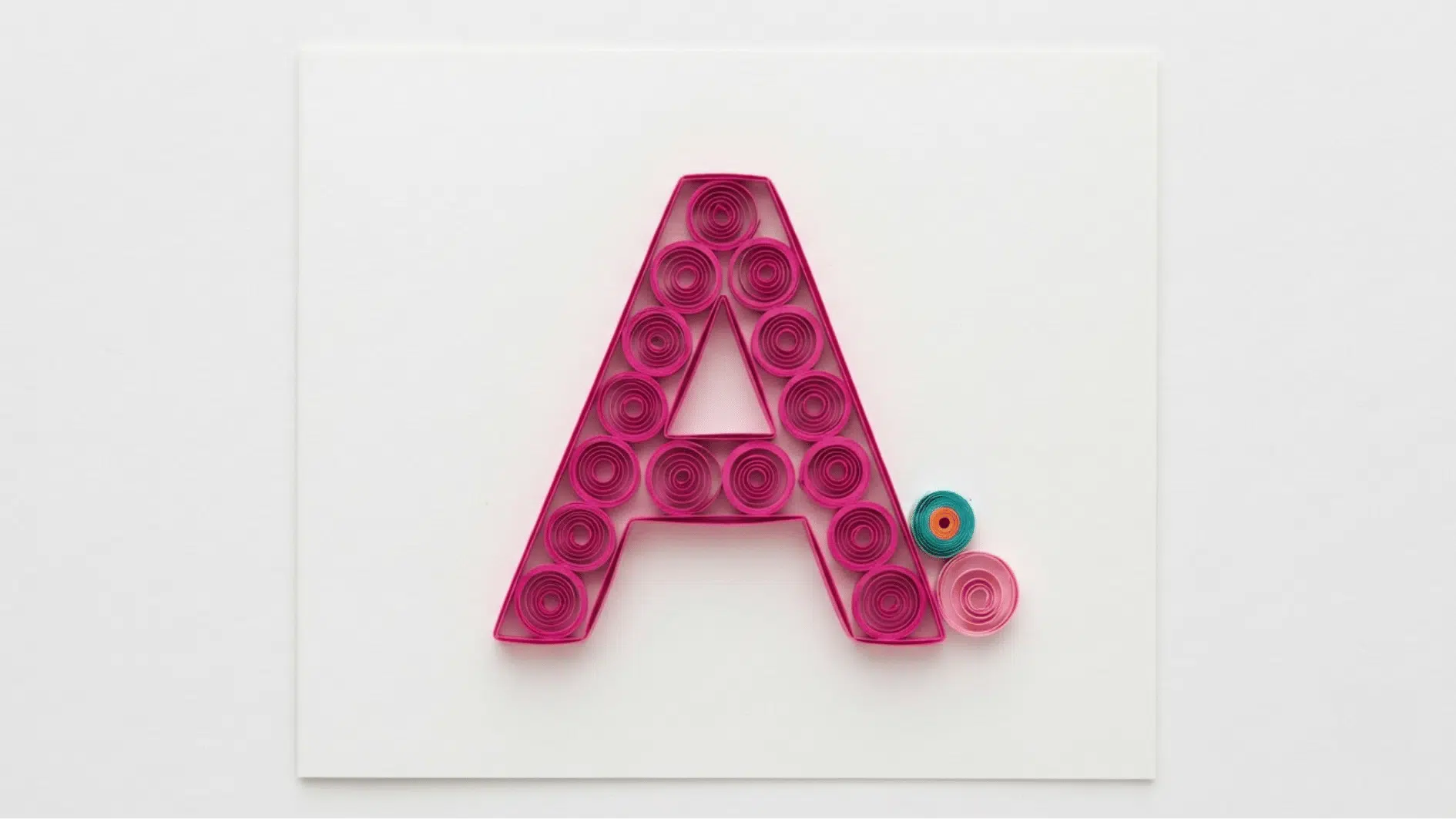 quilled paper letter a in pink coils with small teal and pink accent circles on white background