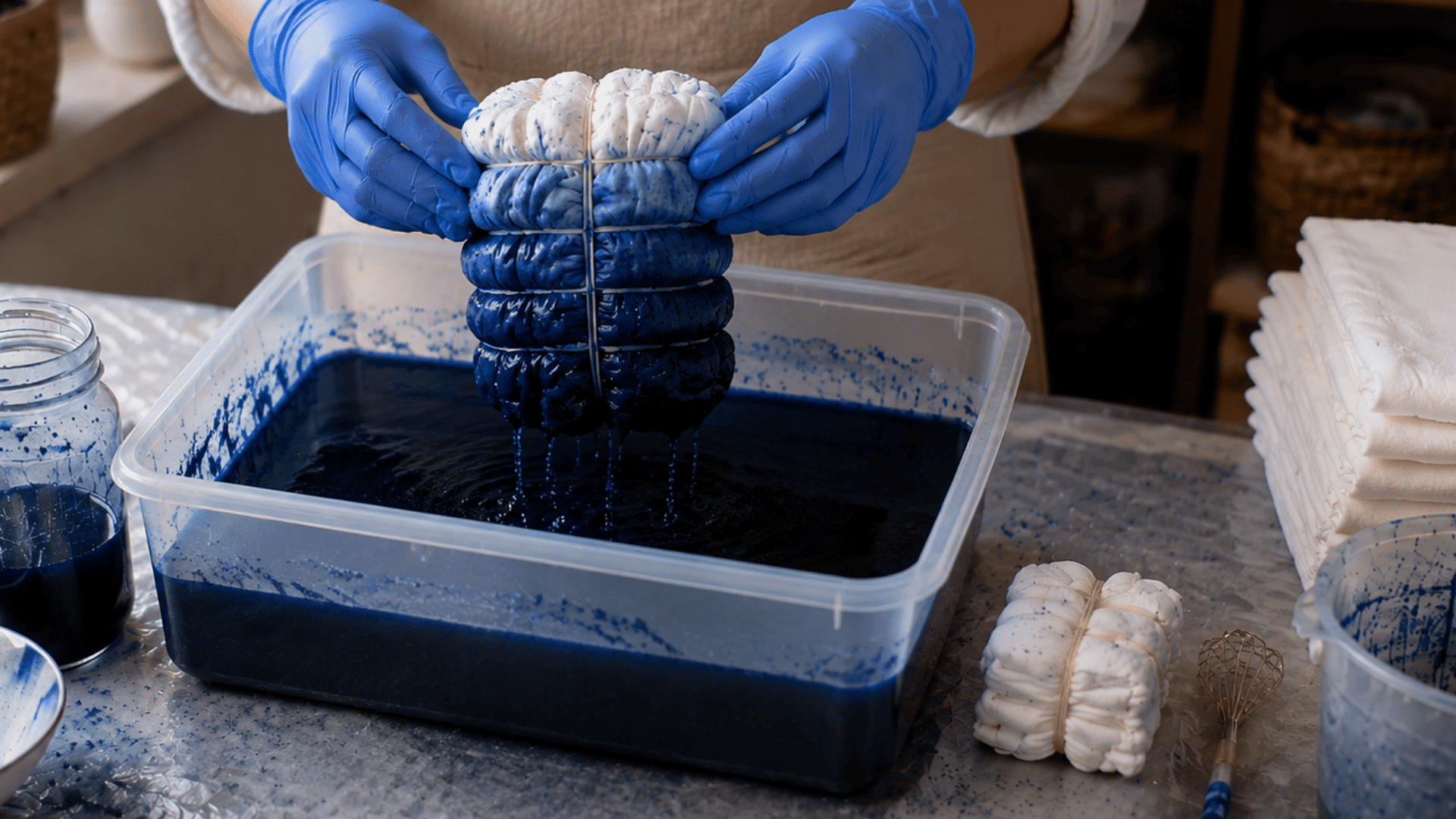 gloved hands lifting dyed shibori fabric bundle with dripping indigo dye into container
