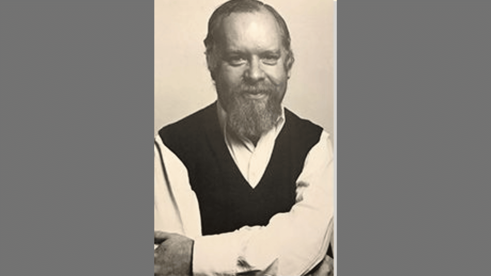 a black-and-white portrait of a smiling bearded man wearing a dark sweater vest over a collared shirt