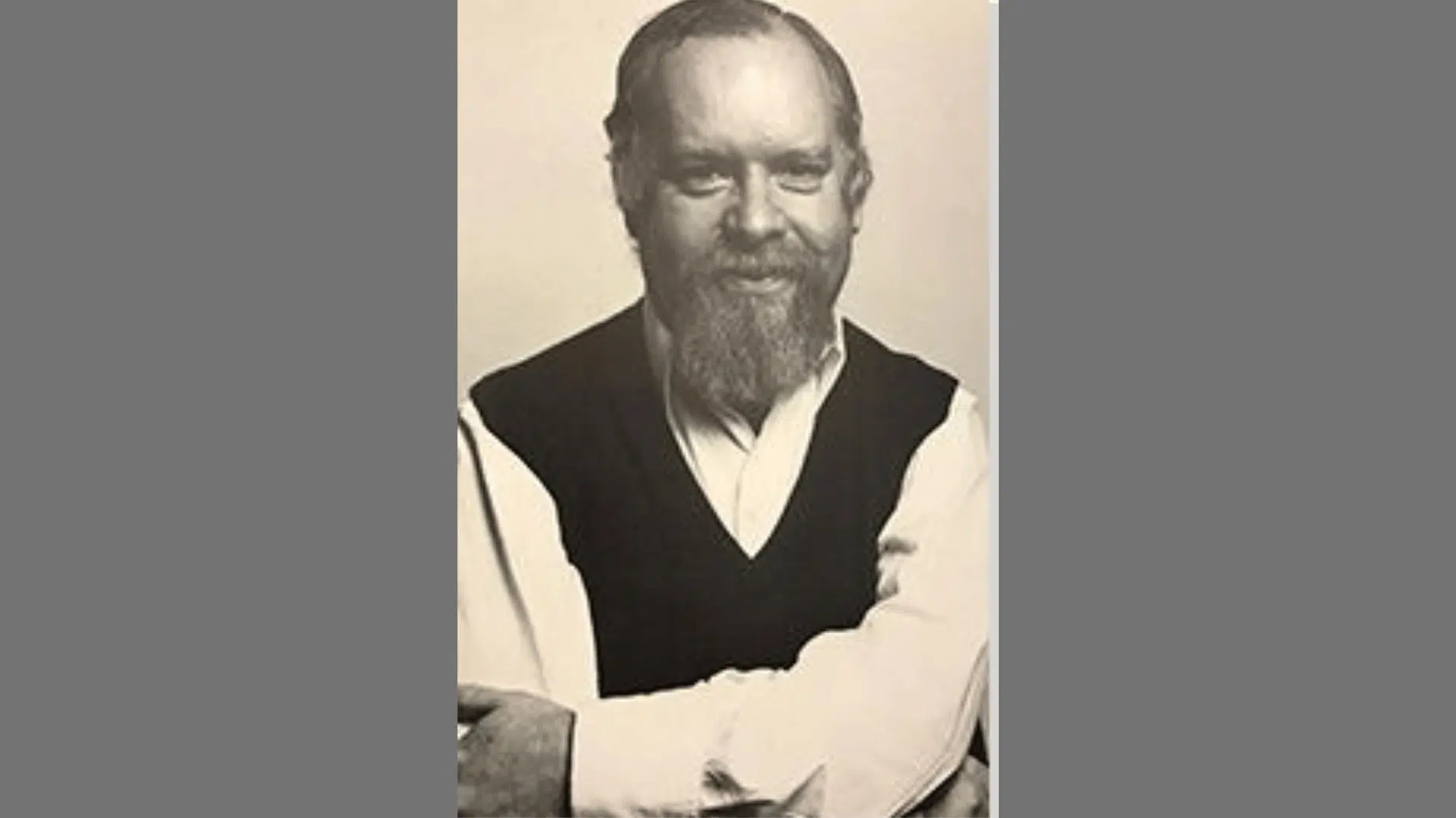 a black-and-white portrait of a smiling bearded man wearing a dark sweater vest over a collared shirt