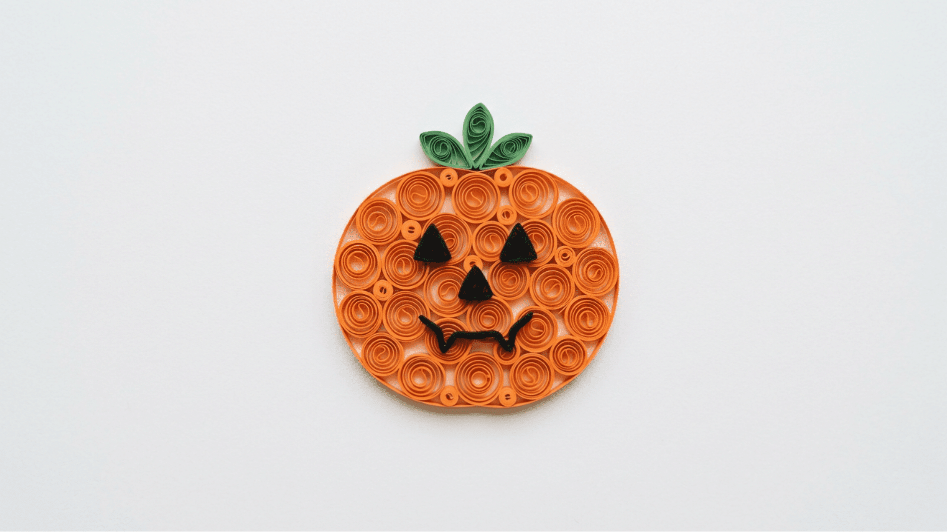quilled paper pumpkin in orange coils with green stem and spooky jack o lantern face on white background