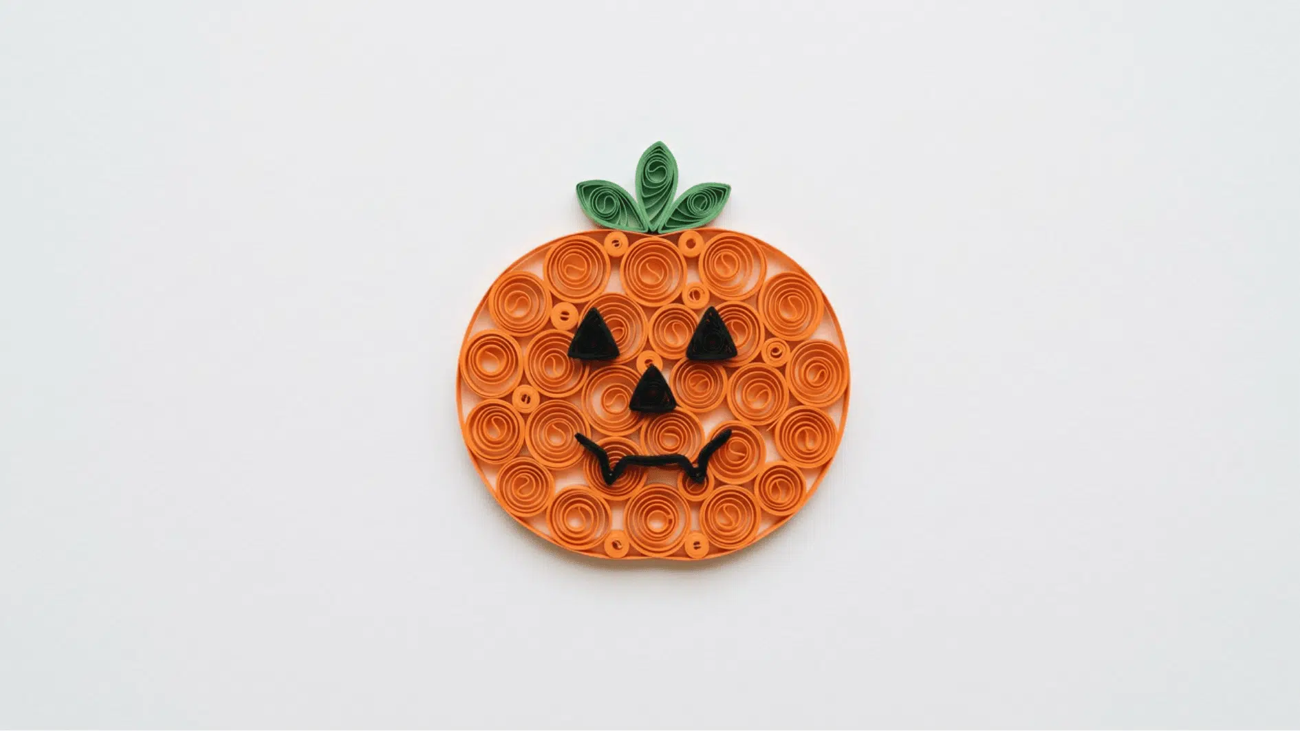 quilled paper pumpkin in orange coils with green stem and spooky jack o lantern face on white background