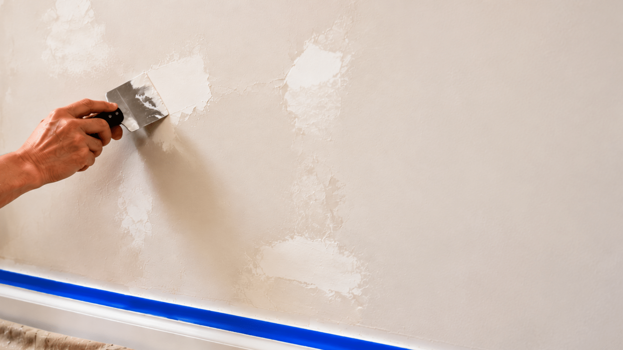 A hand pressing spackle into a wall crack with sandpaper and primer placed nearby on a drop cloth during surface preparation for faux painting.