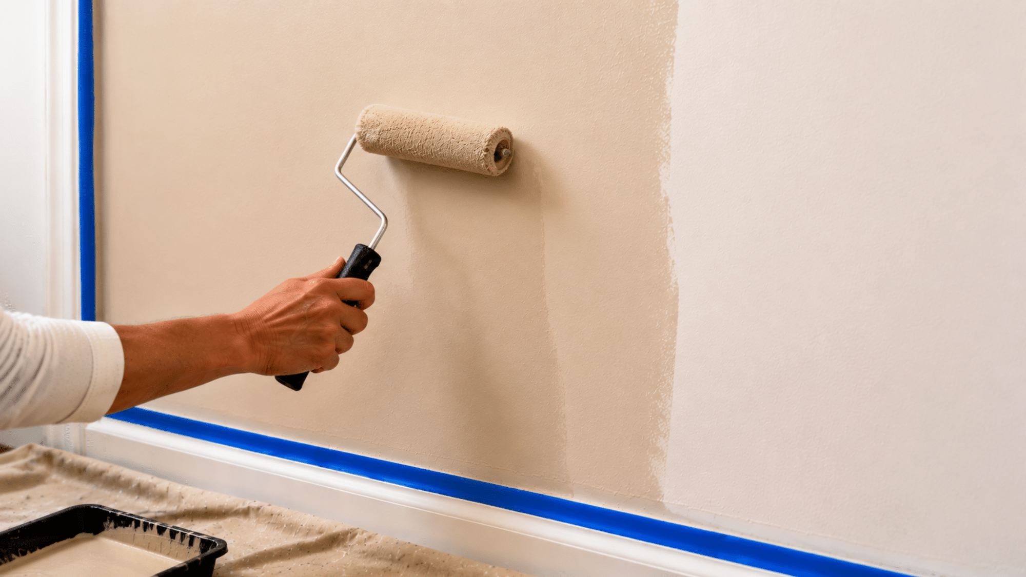 A hand rolling smooth white base coat paint evenly across a freshly primed wall using a paint roller and tray during the faux painting process.