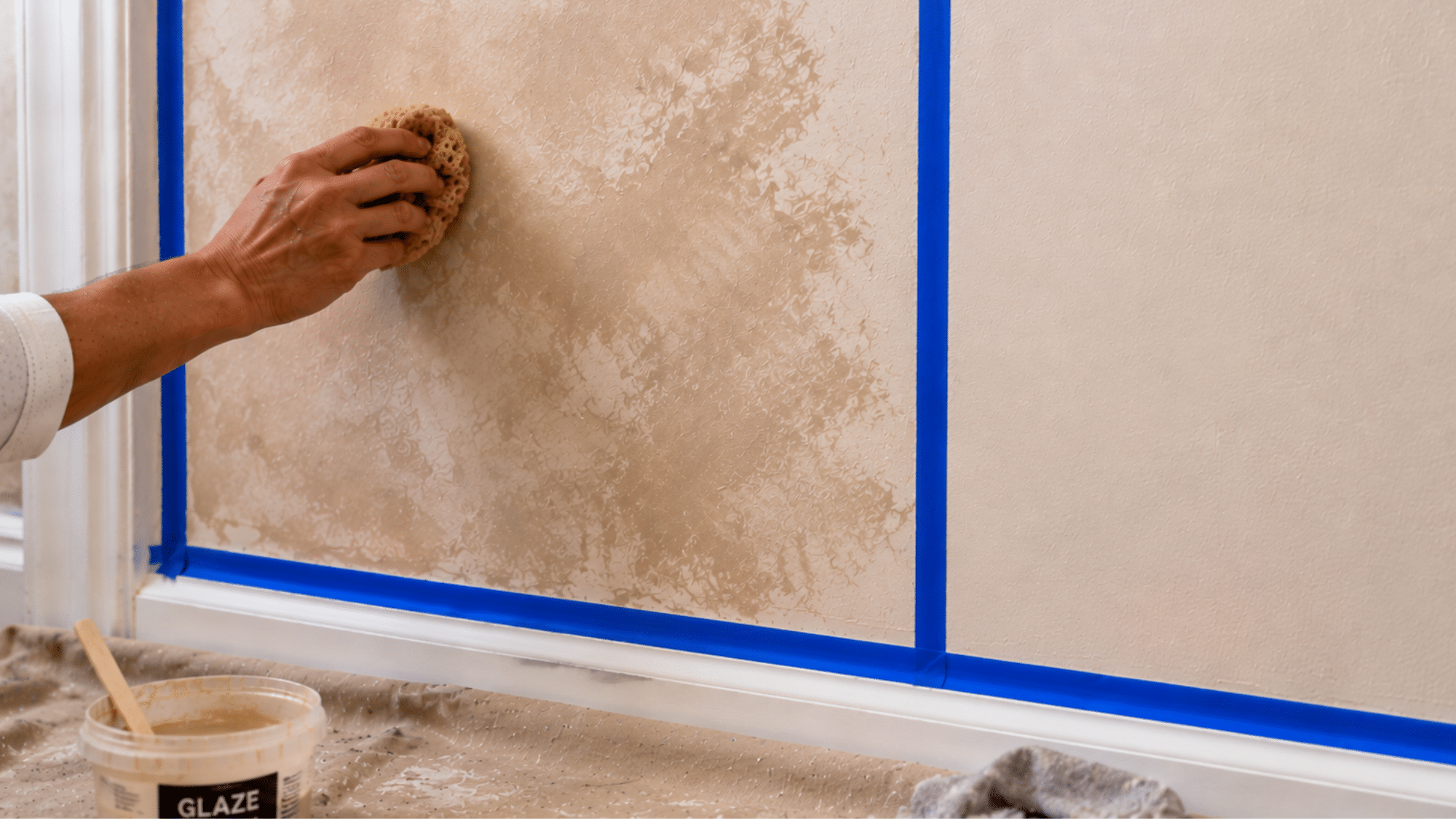 A hand with paint stains pressing a large natural sea sponge loaded with warm golden glaze onto a white wall in small overlapping sections with painter's tape protecting the trim edges