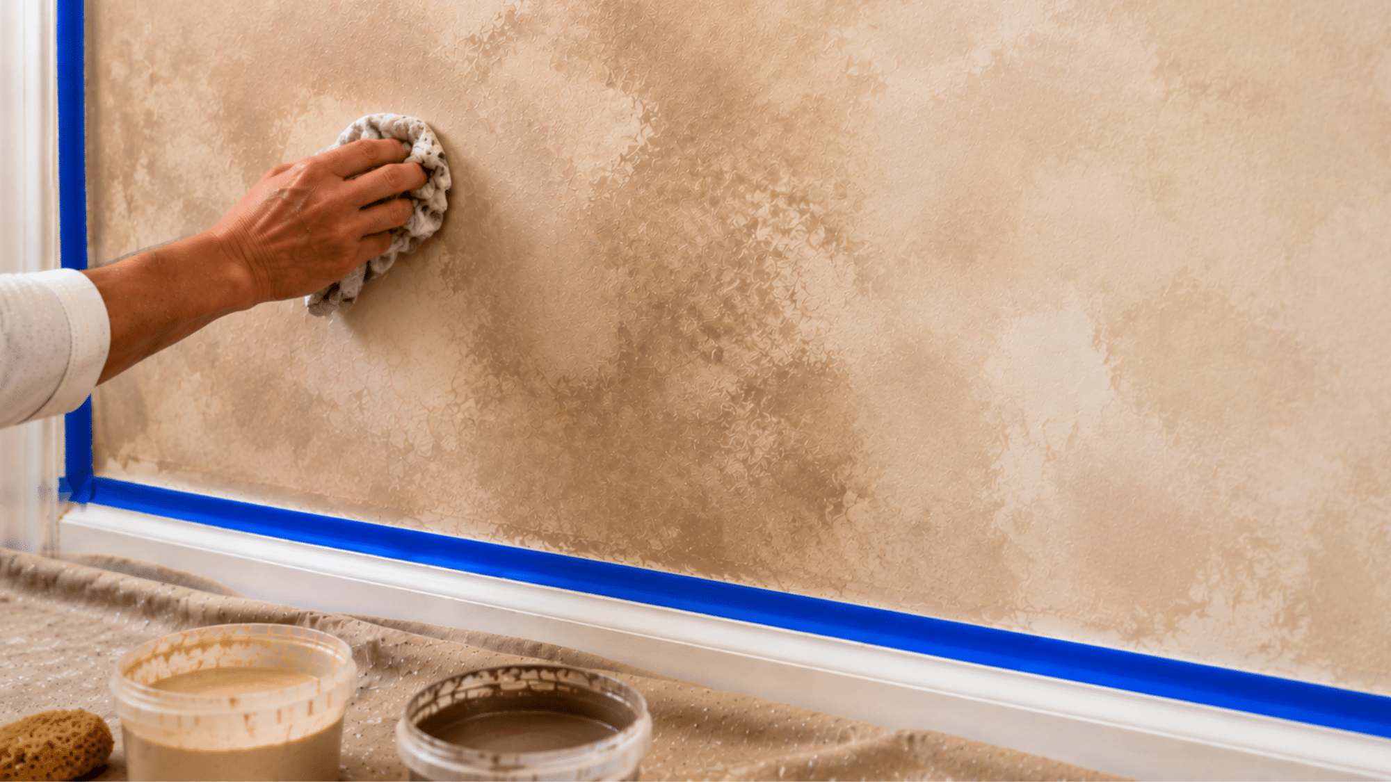 A hand using a wide brush to blend glaze edges on a wall while a second amber glaze layer is being added using a sea sponge resting in a paint tray nearby